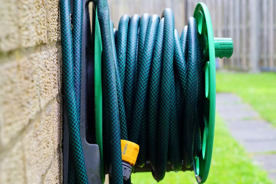 A close-up view of a garden hose coiled neatly on a green plastic reel, mounted on a brick exterior wall. The hose is dark with a blue diamond pattern, appearing flexible and slightly textured, with a